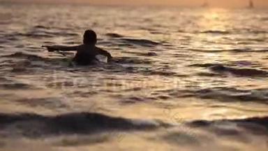 一个男孩<strong>漂浮</strong>在海面上，在<strong>橙色</strong>日落的绝佳背景上坐着冲浪板